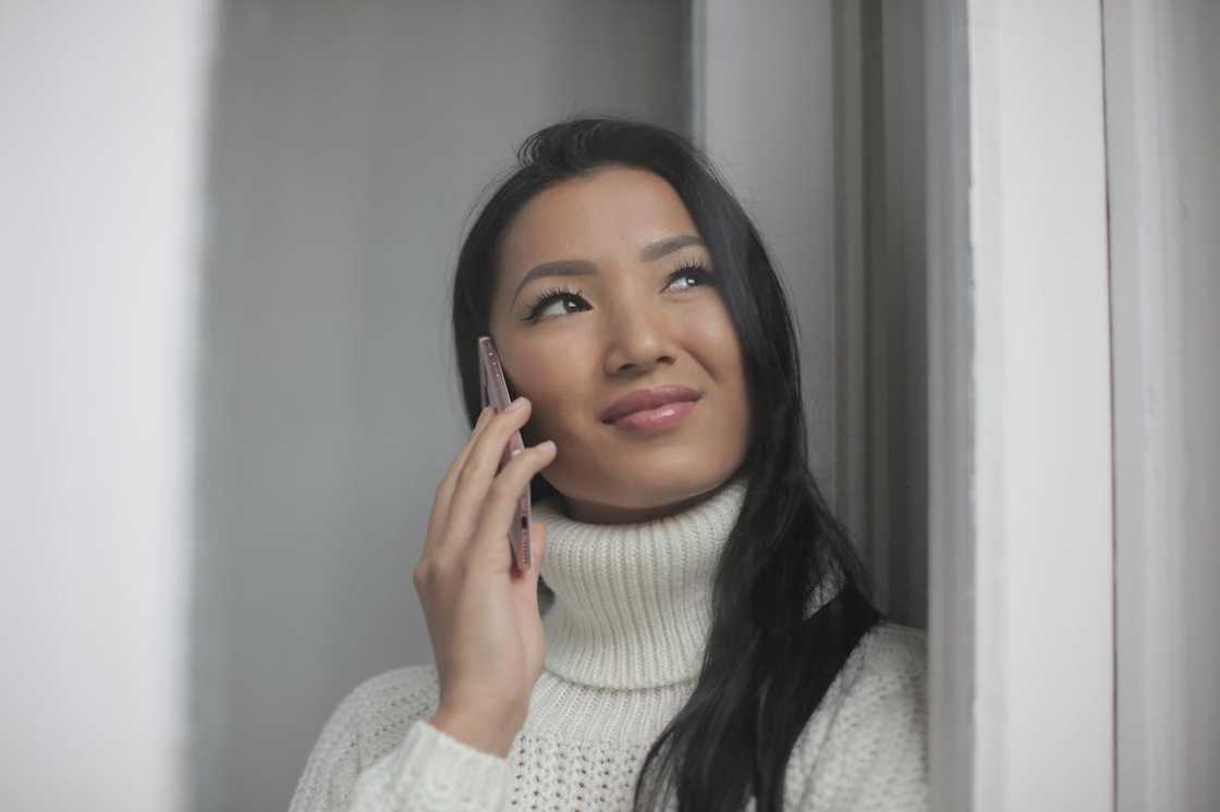 A woman stands by a window holding a phone to her ear, smiling softly as she looks upward. A woman stands by a window holding a phone to her ear, smiling softly as she looks upward.