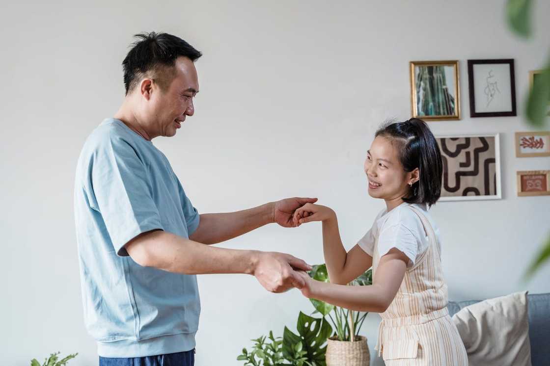 A father and his daughter smile as they hold hands indoors.