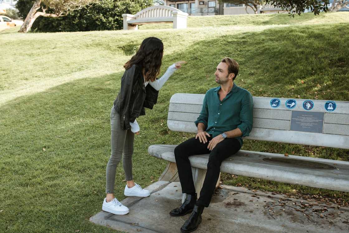 A woman points to a man seated on a bench outdoors.
