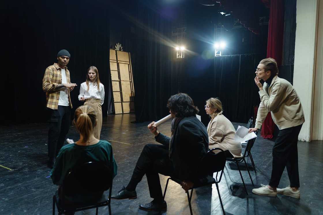 A group of people rehearse on a theatre stage while others sit watching and giving feedback.