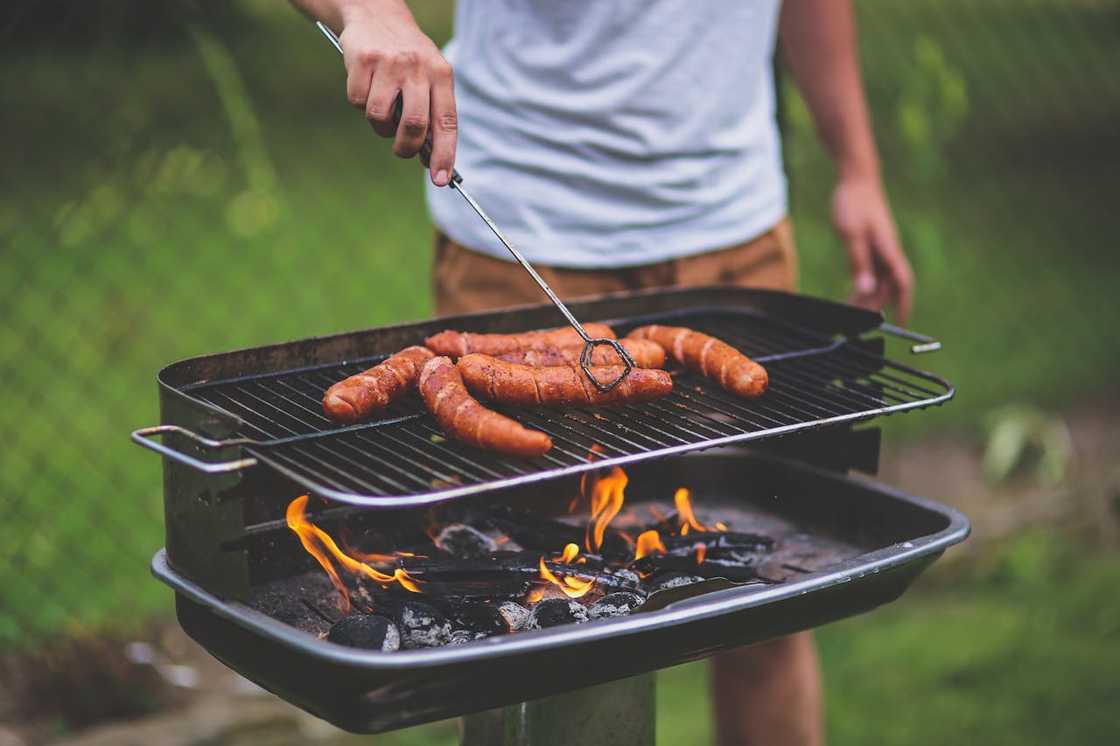 A person grills several sausages over an open flame on an outdoor charcoal barbecue.