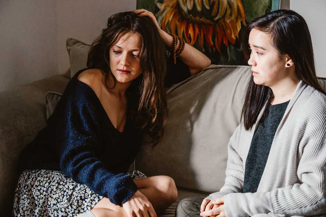 Two women sit on a sofa, one looking distressed while the other listens quietly.