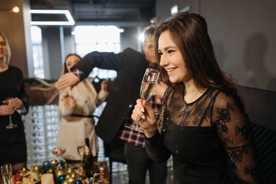 A woman smiling and holding a champagne glass at a party with others in the background. A woman smiling and holding a champagne glass at a party with others in the background.