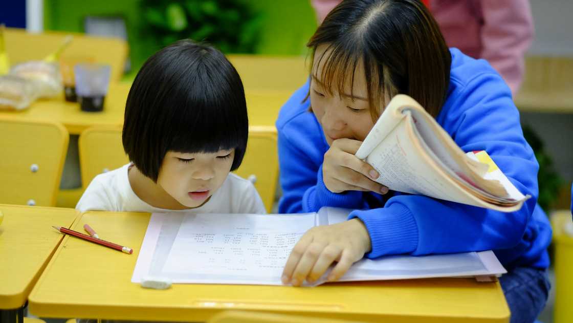 An adult in blue guides a child at a desk with pencils and a worksheet in a classroom.