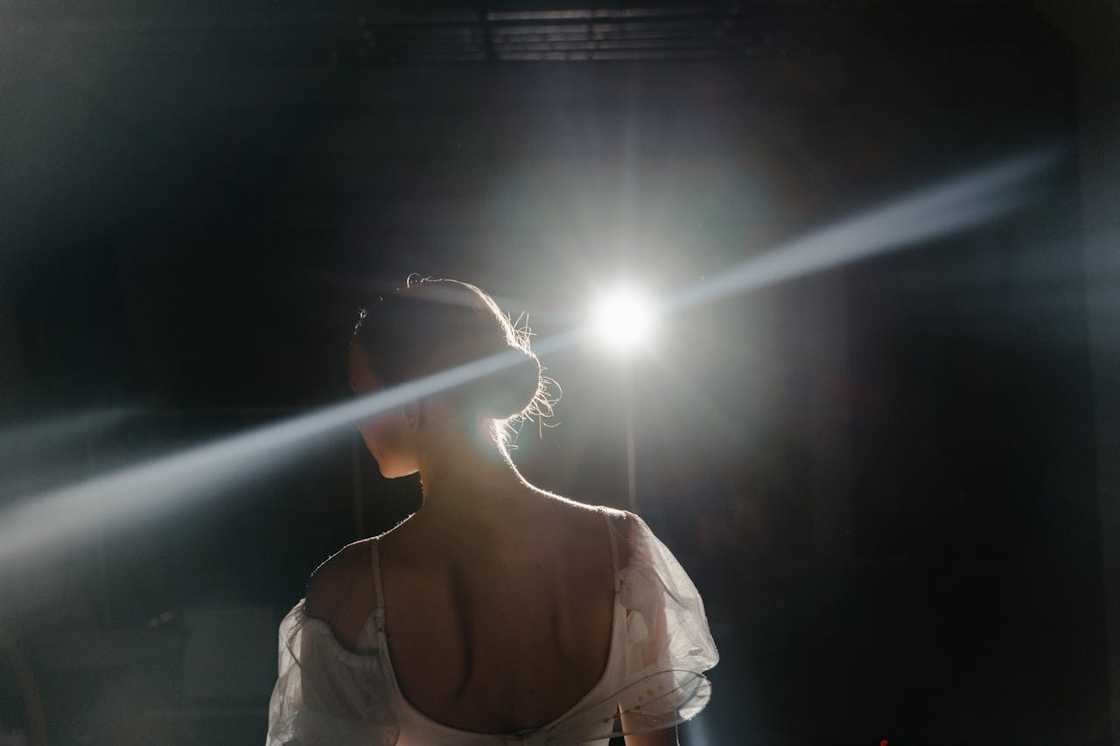 A woman stands on a dark stage with her back to the camera, lit by a bright spotlight.