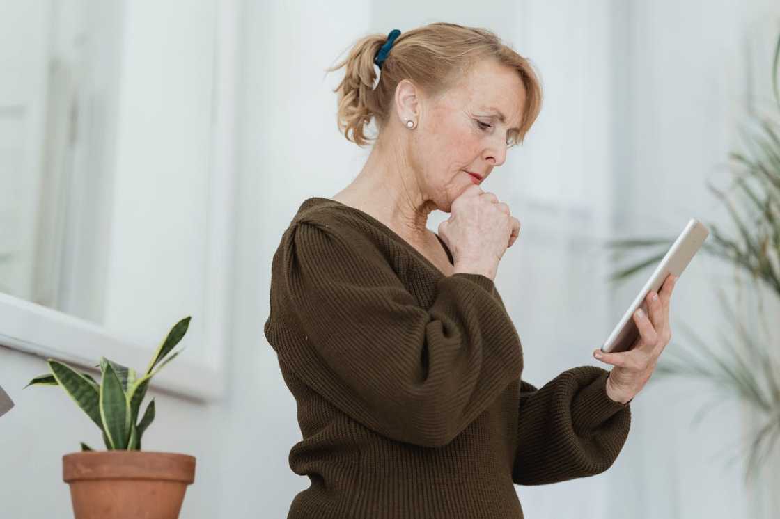 An older woman stands indoors holding a tablet. An older woman stands indoors holding a tablet.
