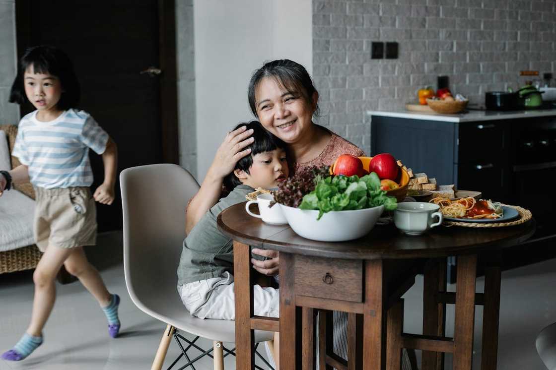 A grandmother hugs her grandson at a kitchen table with food.