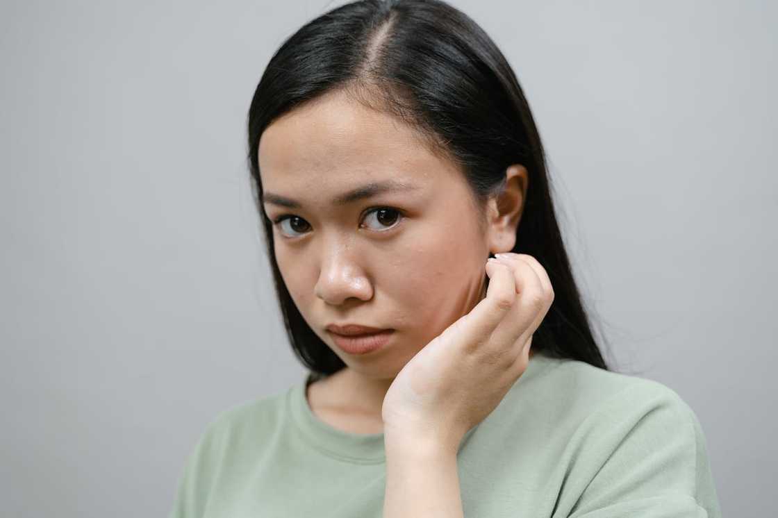 A young woman looks serious while touching her ear against a plain background.