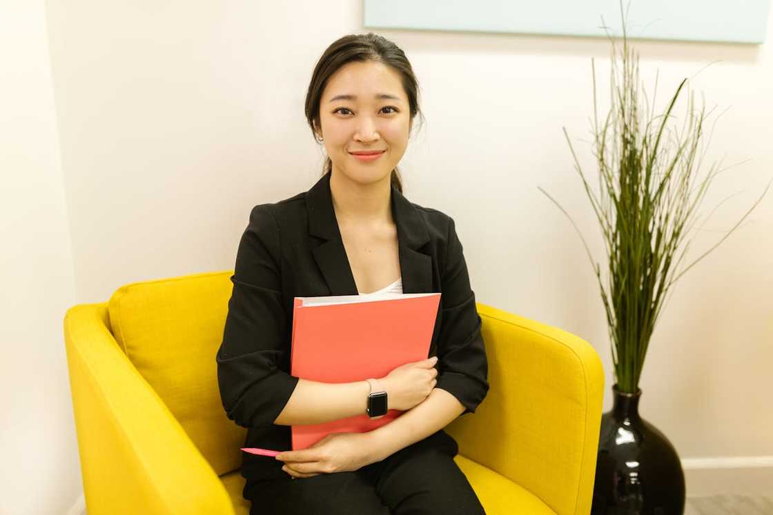 A woman sitting in a yellow chair, holding a folder and smiling. A woman sitting in a yellow chair, holding a folder and smiling.