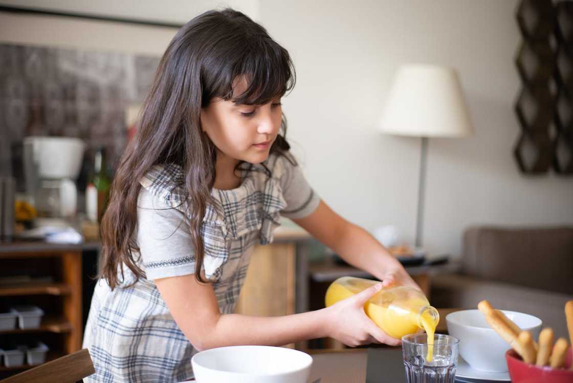 A young girl pours orange juice into a glass at a dining table.