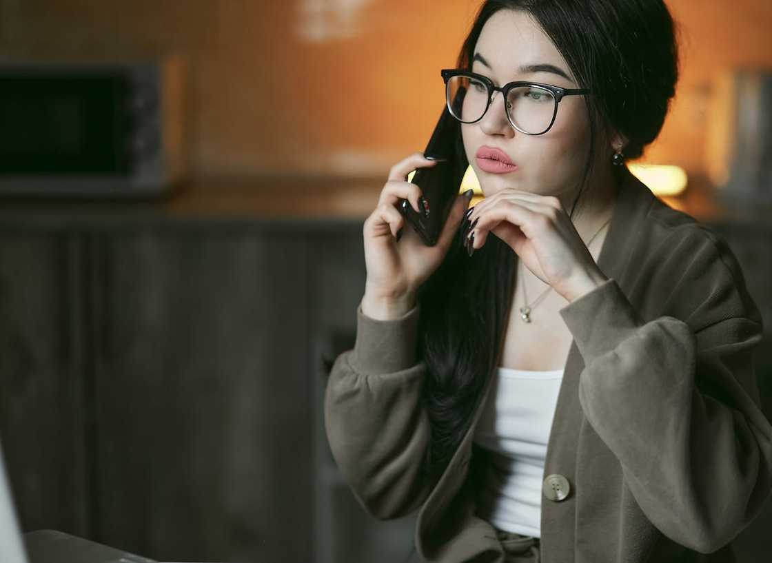 A young woman wearing glasses talks on a phone indoors with a focused expression. A young woman wearing glasses talks on a phone indoors with a focused expression.