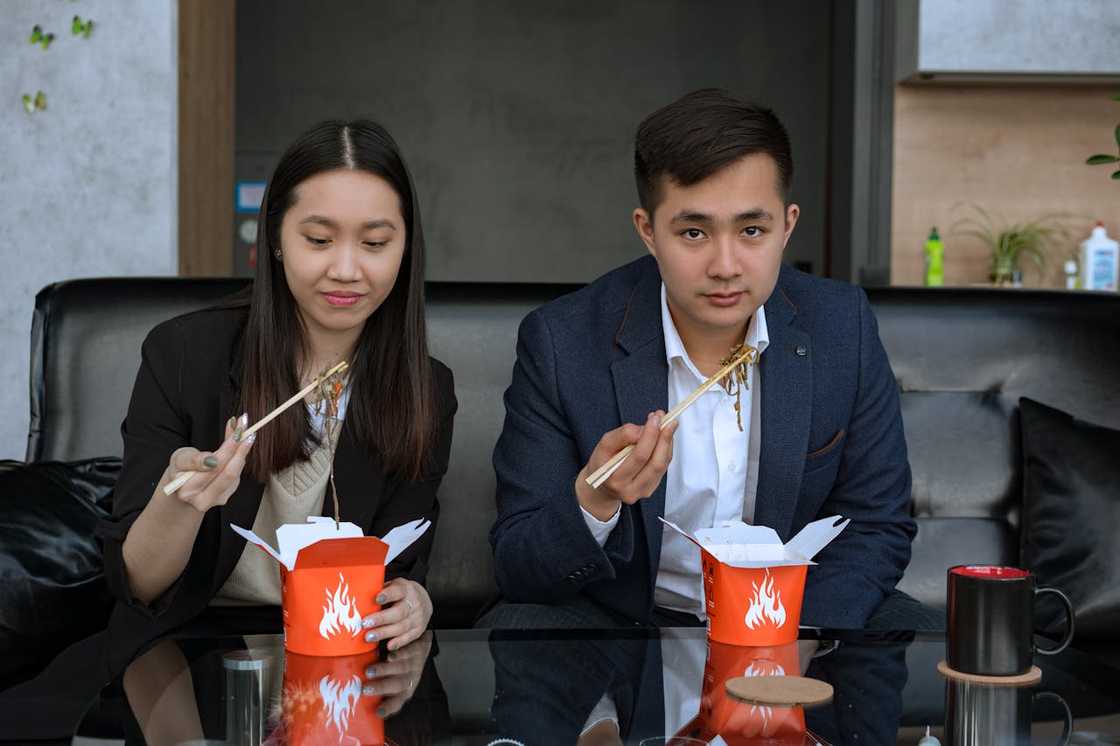 Two co-workers eat noodles from orange takeout boxes with chopsticks.