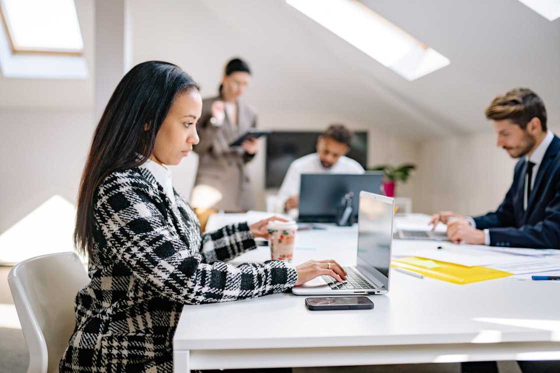 A woman working on a laptop at a shared office table while colleagues appear in the background. A woman working on a laptop at a shared office table while colleagues appear in the background.