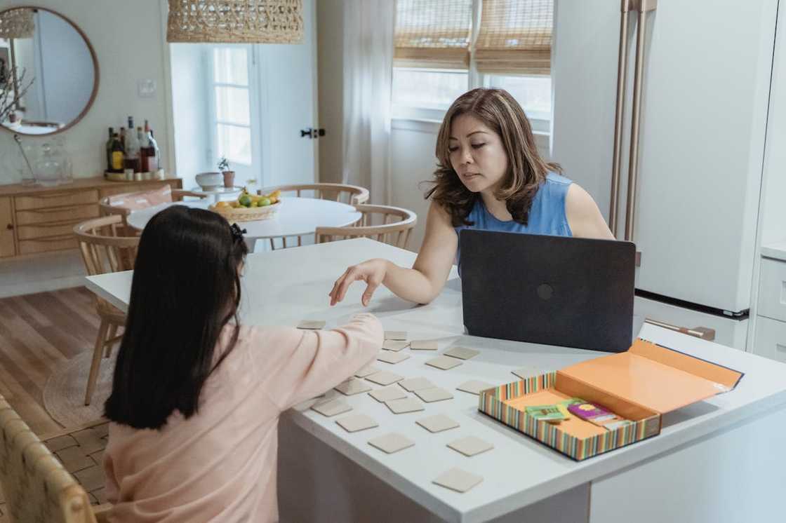 Adult and child sit at a table with a laptop, working together as city lights glow outside. Adult and child sit at a table with a laptop, working together as city lights glow outside.