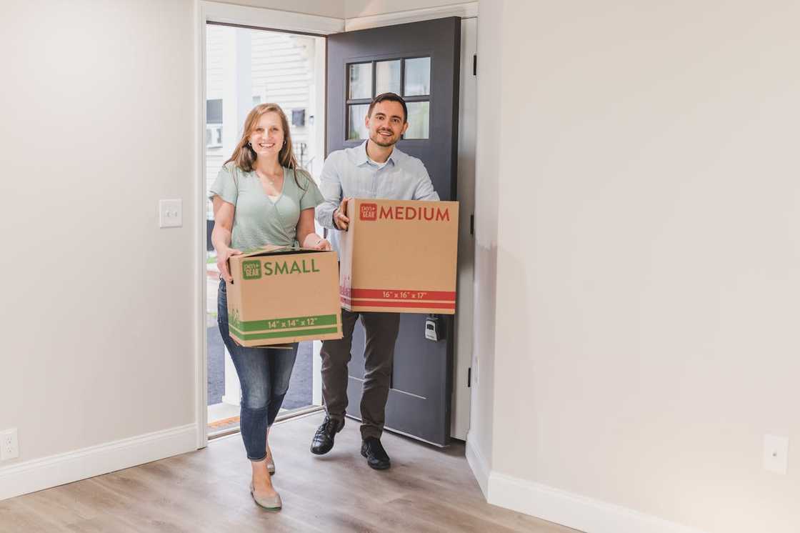 A smiling couple carrying moving boxes into a new home through the front door.