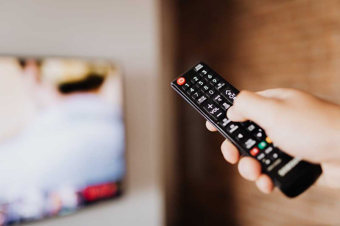 A hand holds a TV remote control pointed toward a blurred television screen.