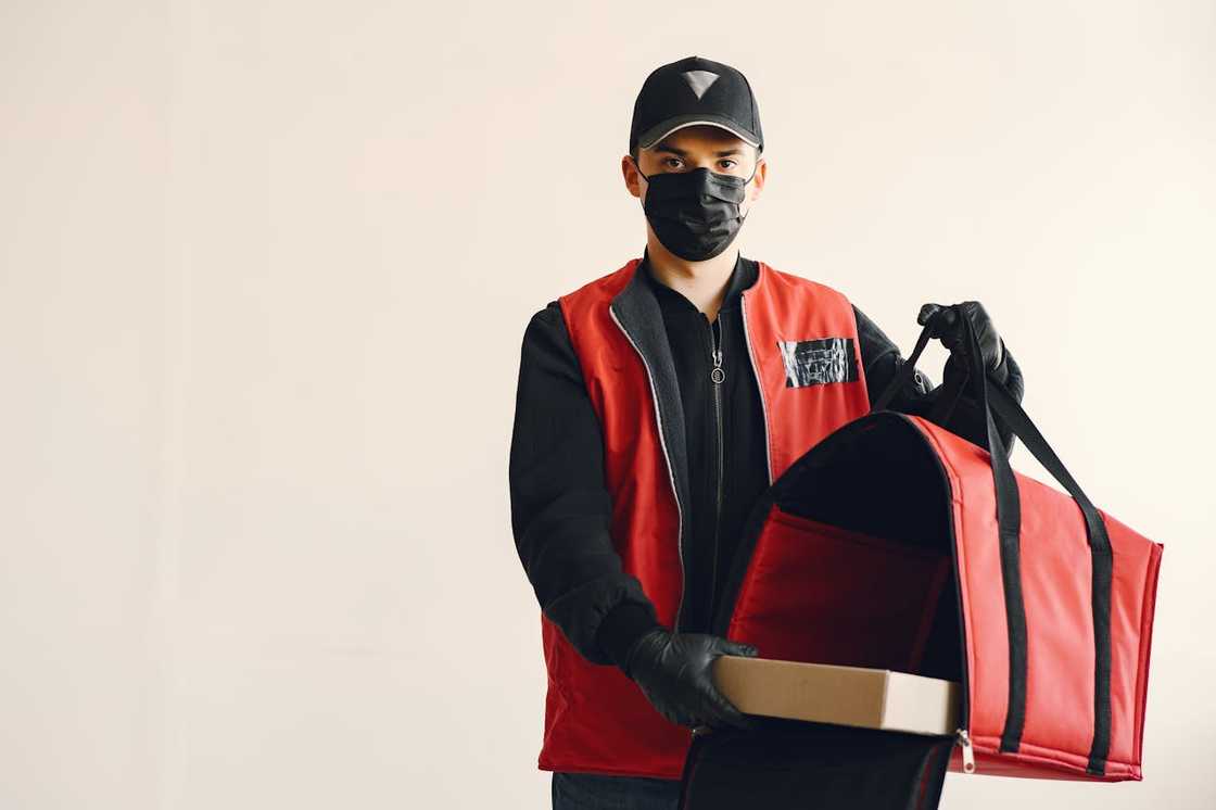 A delivery worker wearing a mask holding a food delivery bag and box. A delivery worker wearing a mask holding a food delivery bag and box.