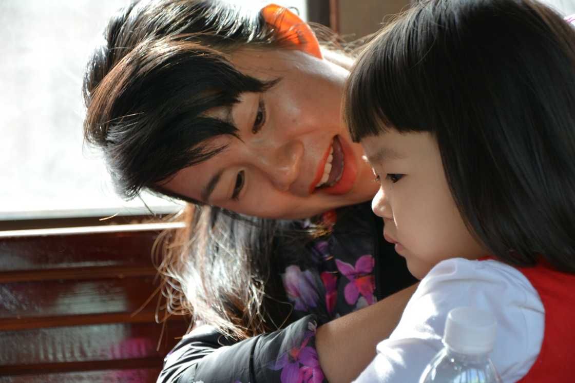 An adult in a floral jacket leans in toward a child in a red vest during a quiet moment indoors.