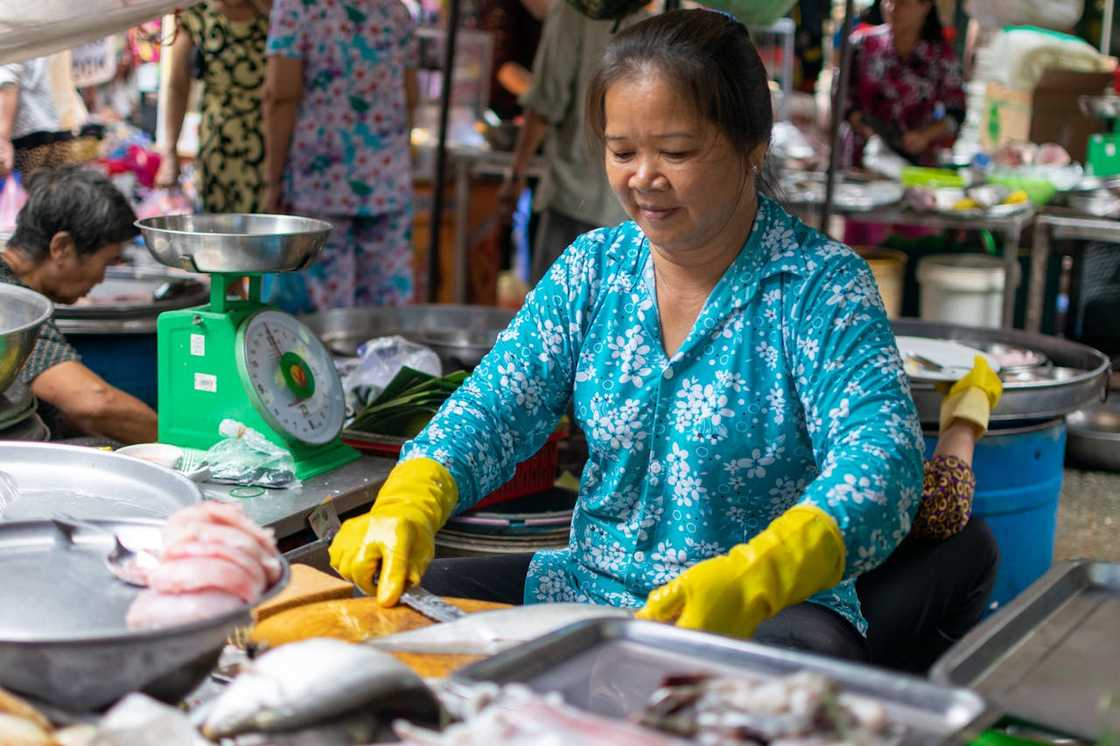 A woman wearing yellow gloves preparing fish at a busy open-air market stall. A woman wearing yellow gloves preparing fish at a busy open-air market stall.