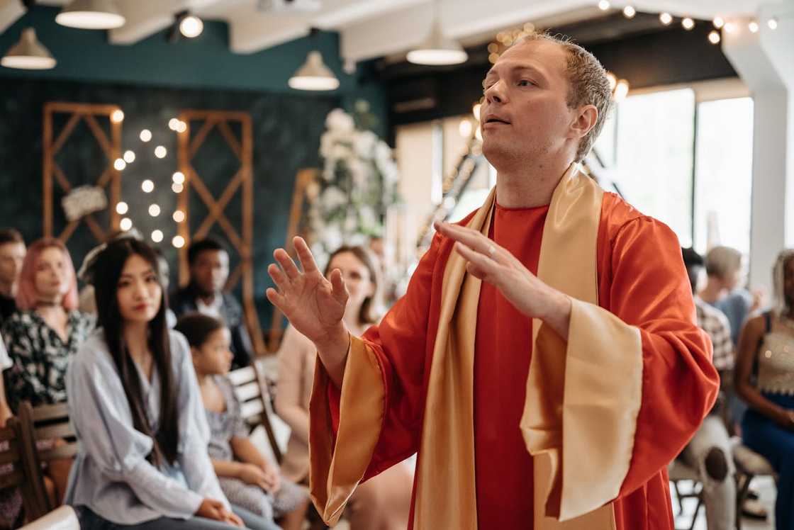 A man in ceremonial robes speaking with raised hands before a seated audience indoors.