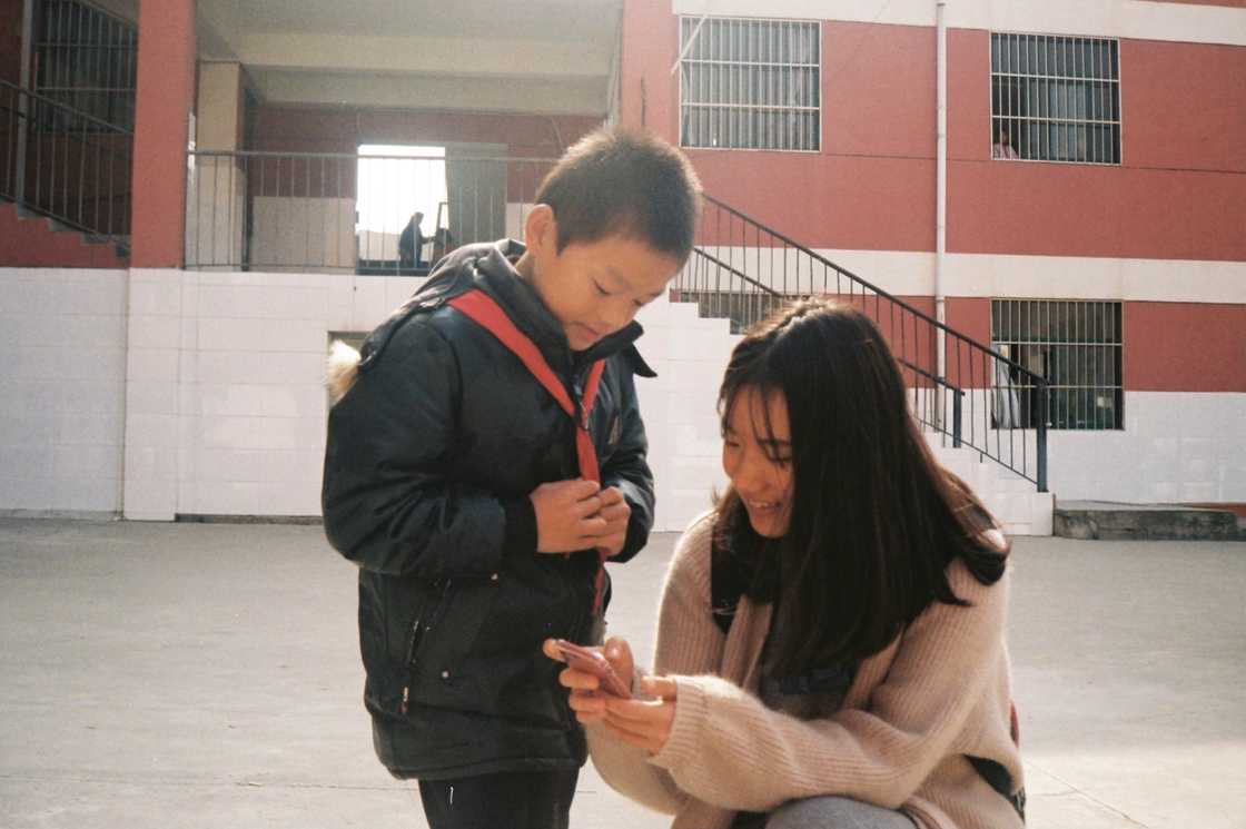 A child in a winter coat looks at a smartphone held by a smiling adult kneeling beside them.