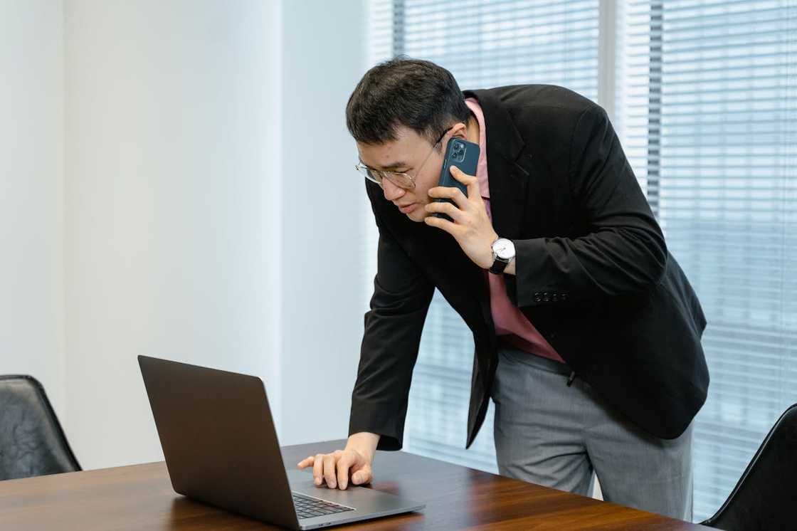A man leans over a table, talking on his phone while looking at a laptop screen.