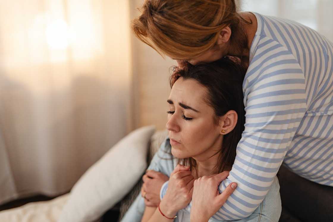 A woman with closed eyes sits looking distressed while another person embraces her from behind. A woman with closed eyes sits looking distressed while another person embraces her from behind.