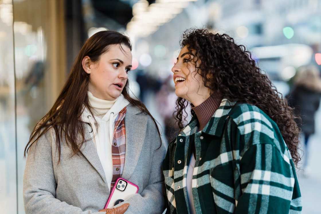 Two women stand close together and talk on a busy street.