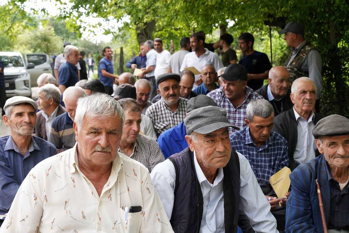 A large group of elderly men is seated outdoors under trees, listening attentively. A large group of elderly men is seated outdoors under trees, listening attentively.