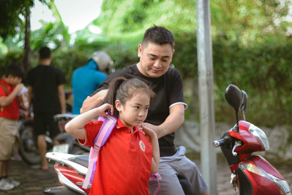 A man helps a girl in a red school shirt put on a backpack near a motorbike outdoors.