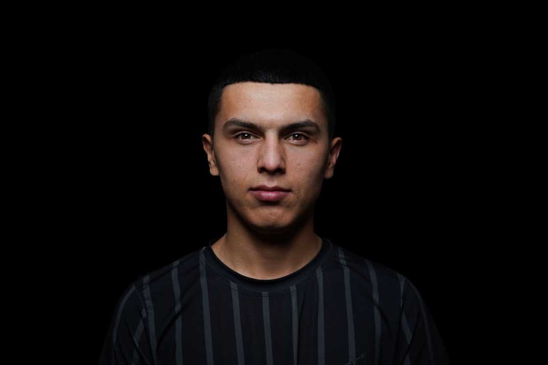 A young man looks directly at the camera against a dark background.