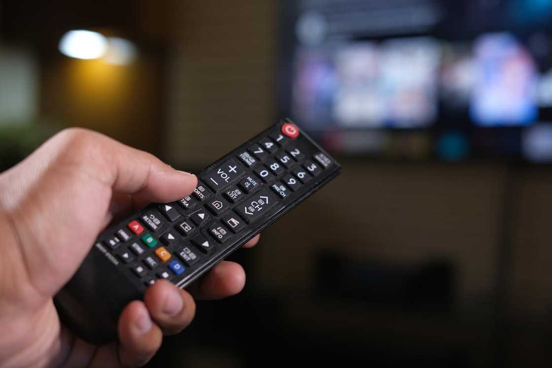 A hand holds a TV remote control pointed toward a blurred television screen.
