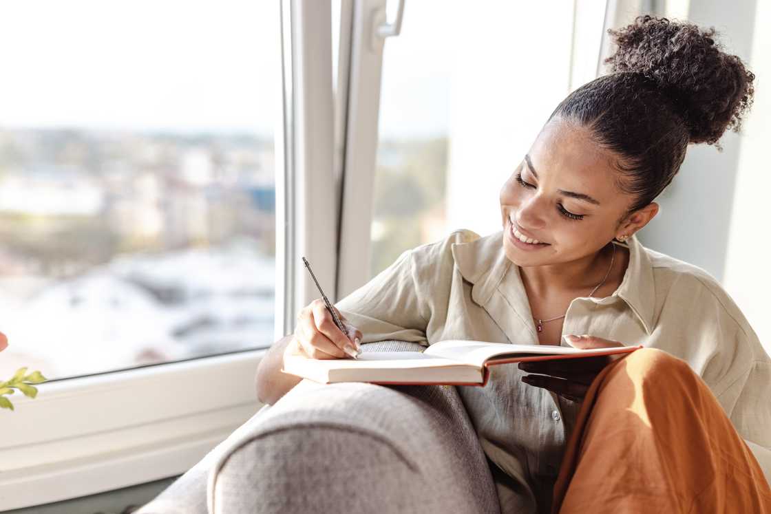 Person sits on a couch near a window, smiling while writing in a notebook. Person sits on a couch near a window, smiling while writing in a notebook.