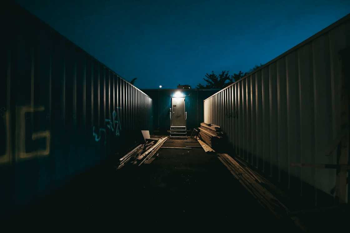A narrow walkway between shipping containers leads to a lit door at night. A narrow walkway between shipping containers leads to a lit door at night.