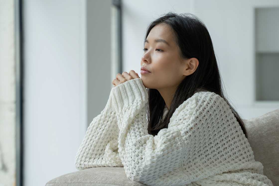 A serious young woman sitting on the couch looking out the window
