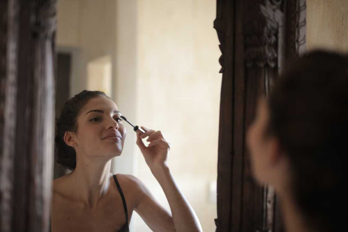 A woman applies mascara while looking at herself in a mirror.