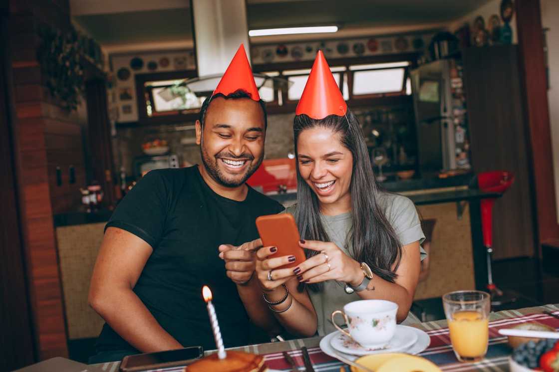 Two people wearing party hats sit at a table, smiling as they look at a smartphone beside a lit birthday candle.
