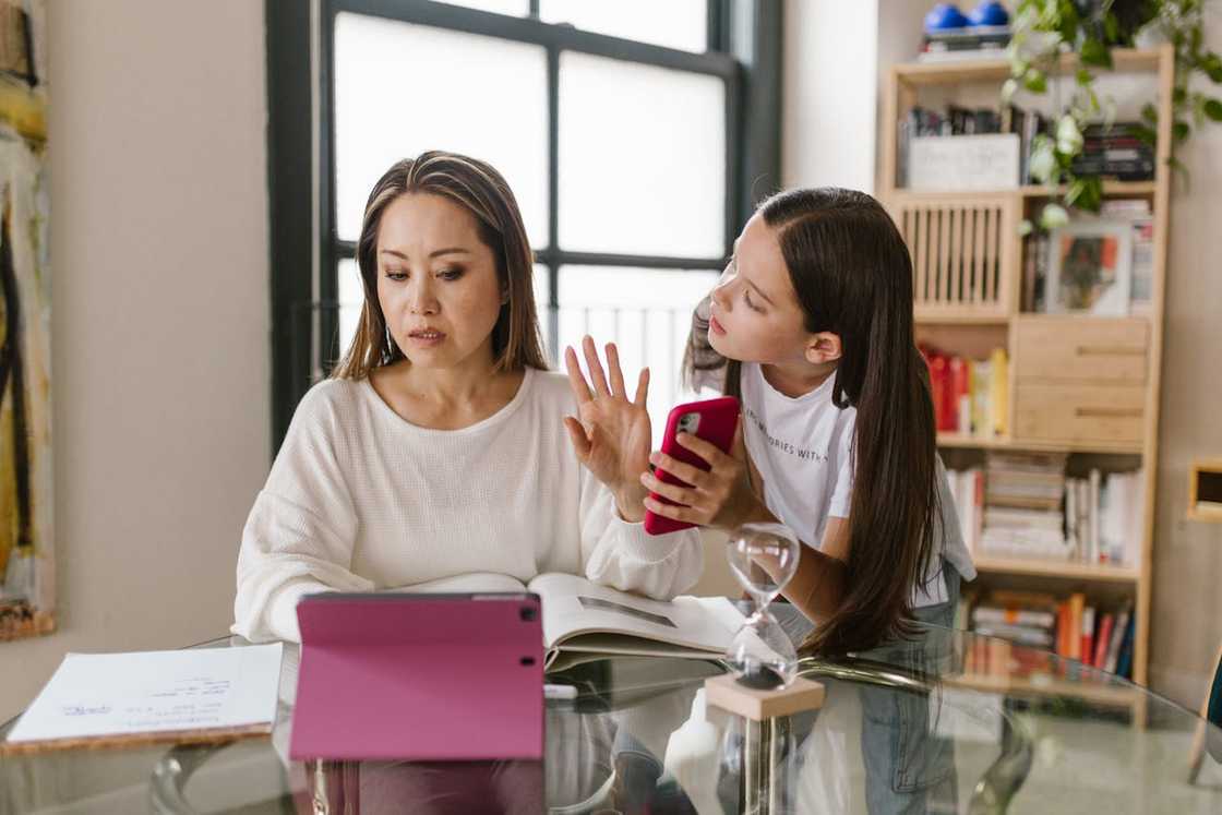 A person raises a hand at a table with a tablet, book, and notes, while another holds a phone.