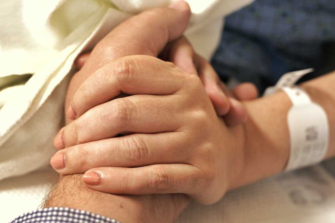 Older person lies in a hospital bed while another individual gently holds their hand in a comforting gesture. Older person lies in a hospital bed while another individual gently holds their hand in a comforting gesture.