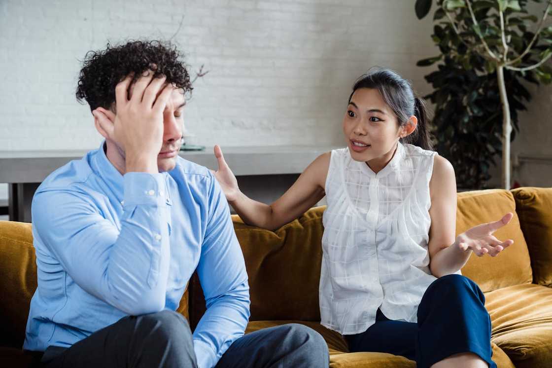 A woman speaks animatedly to a distressed man sitting on a couch.