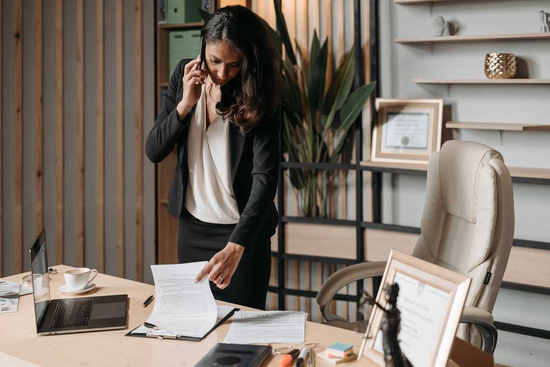 A female lawyer is talking on the phone while reviewing documents at an office desk. A female lawyer is talking on the phone while reviewing documents at an office desk.