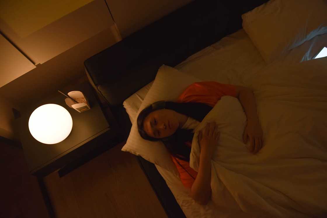 A woman lies awake in bed at night beside a glowing bedside lamp.