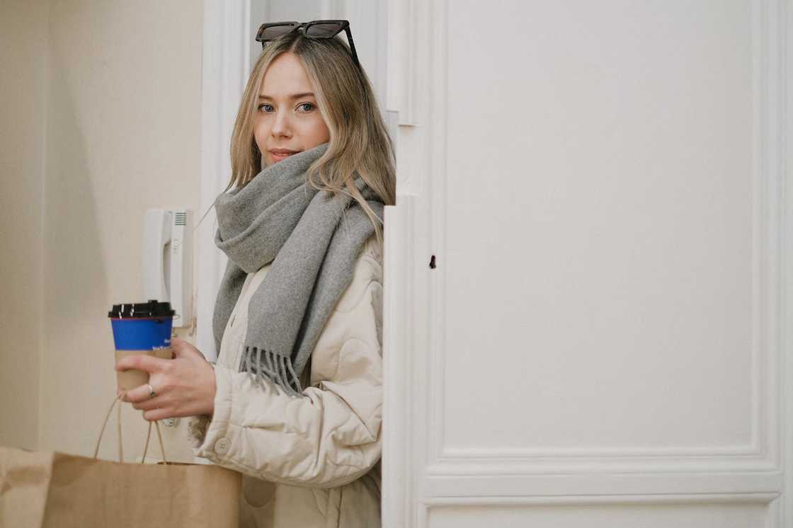A woman stands in a doorway holding a coffee cup and a shopping bag, looking back calmly. A woman stands in a doorway holding a coffee cup and a shopping bag, looking back calmly.