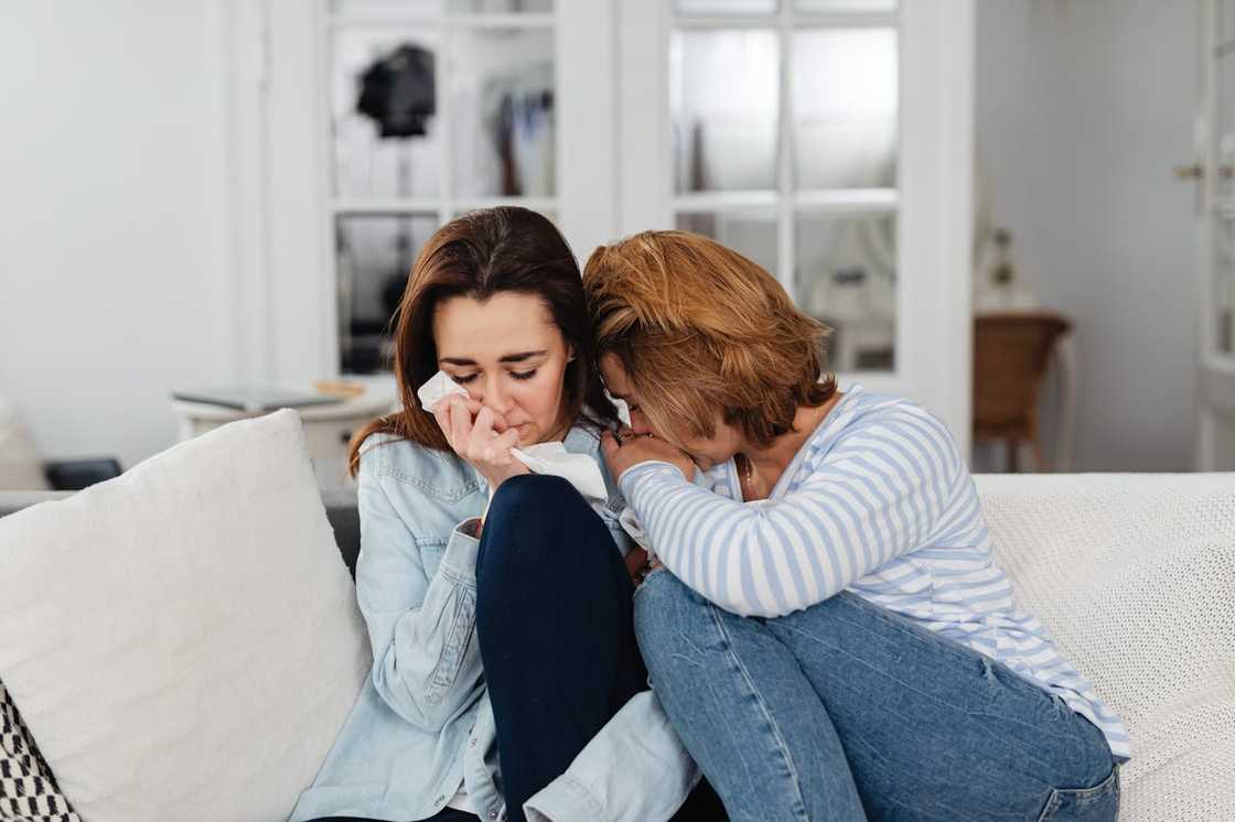 Two women sit close together on a couch crying together.