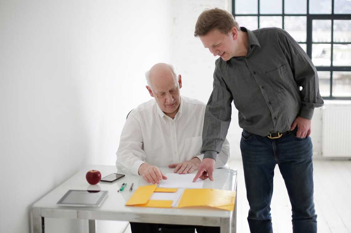 A younger man points at documents while speaking with an older man at a desk. A younger man points at documents while speaking with an older man at a desk.