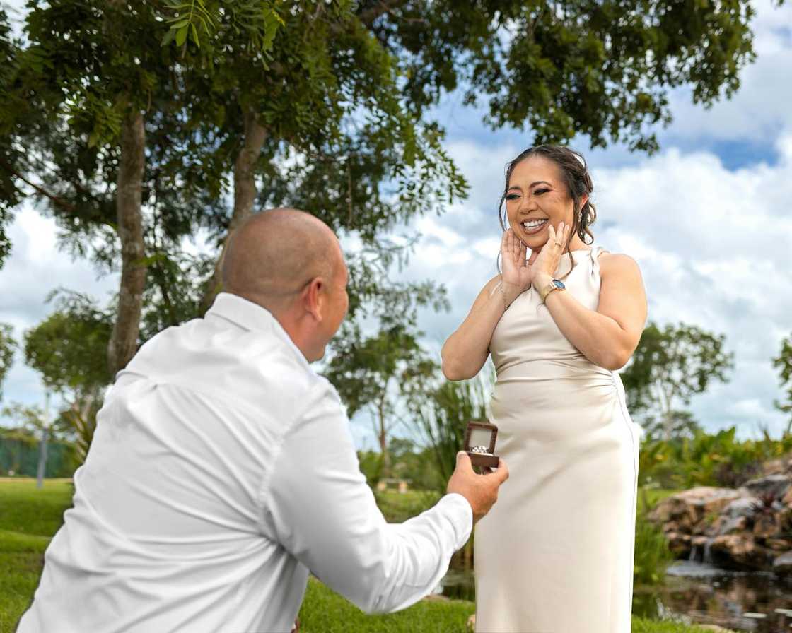 Man kneeling and proposing with a ring to a smiling woman outdoors in a park.