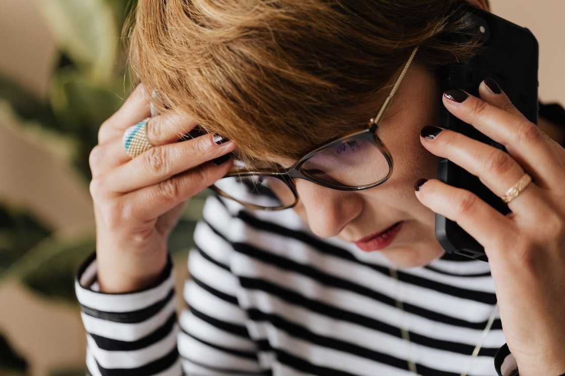 A woman wearing glasses holds a phone to her ear appearing stressed during a call.