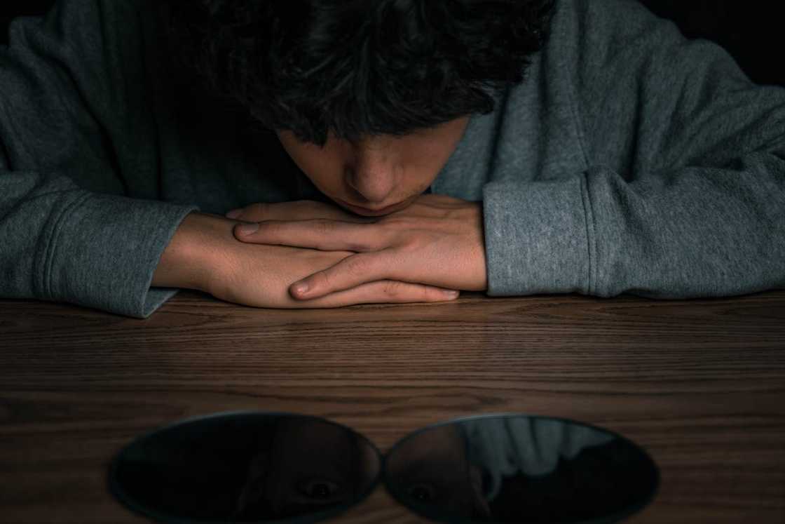A distressed young Asian man rests his head on his hands on a table.
