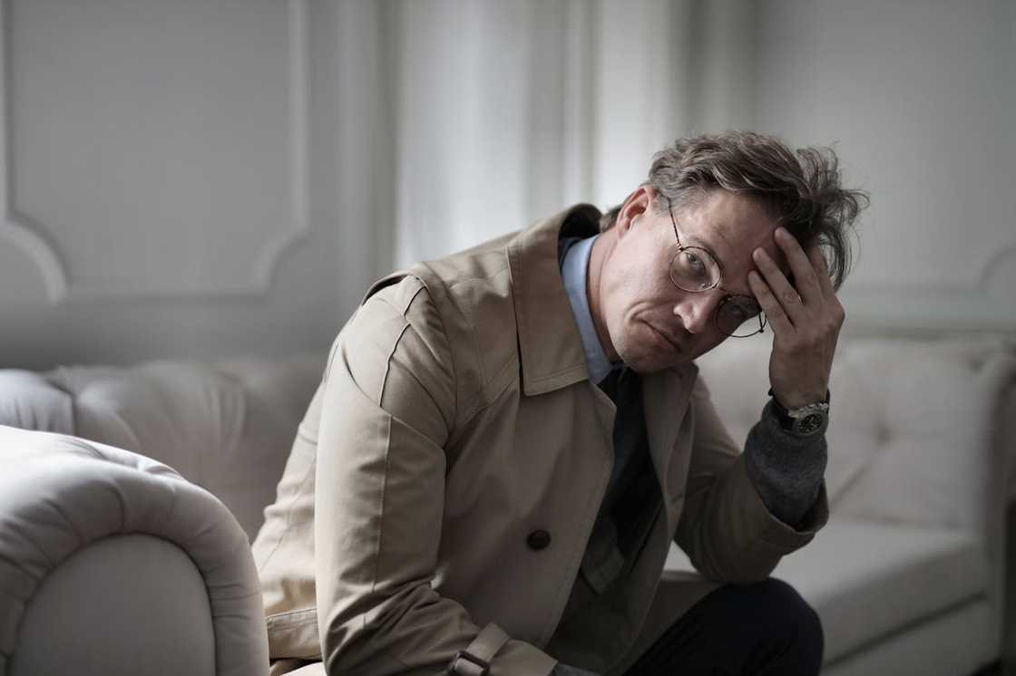 A man wearing glasses sits on a sofa, leaning forward with his hand on his forehead, looking distressed. A man wearing glasses sits on a sofa, leaning forward with his hand on his forehead, looking distressed.