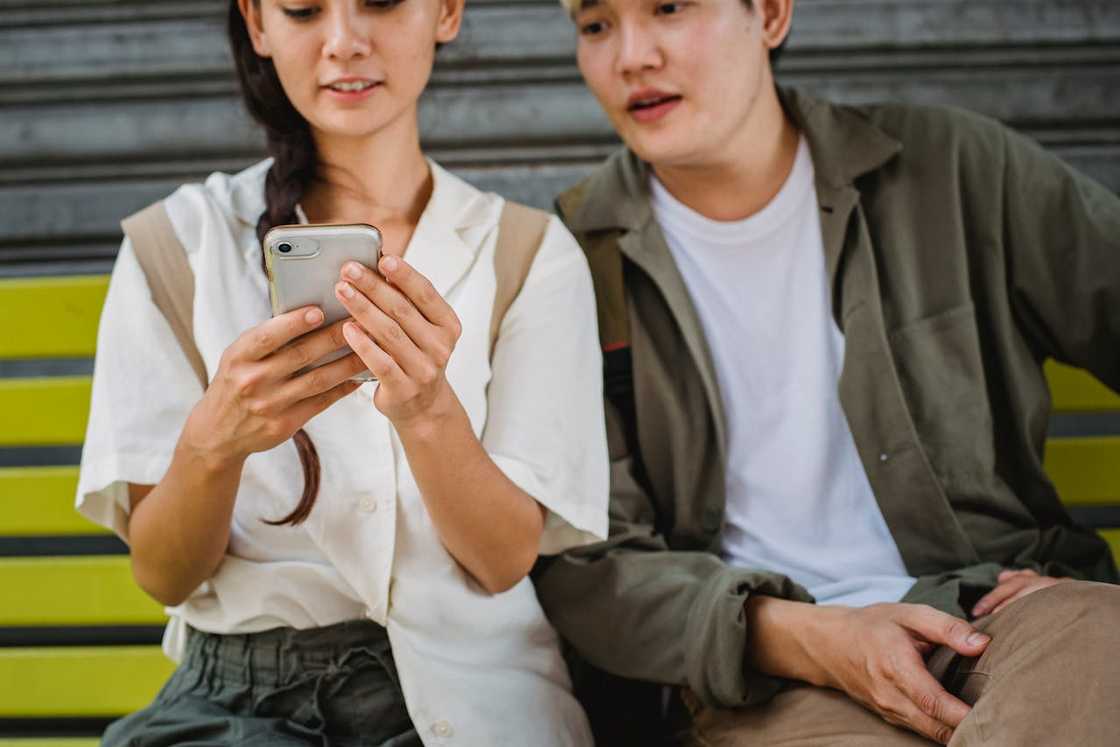 A woman sits on a bench holding a smartphone while a man beside her leans in to look at the screen.